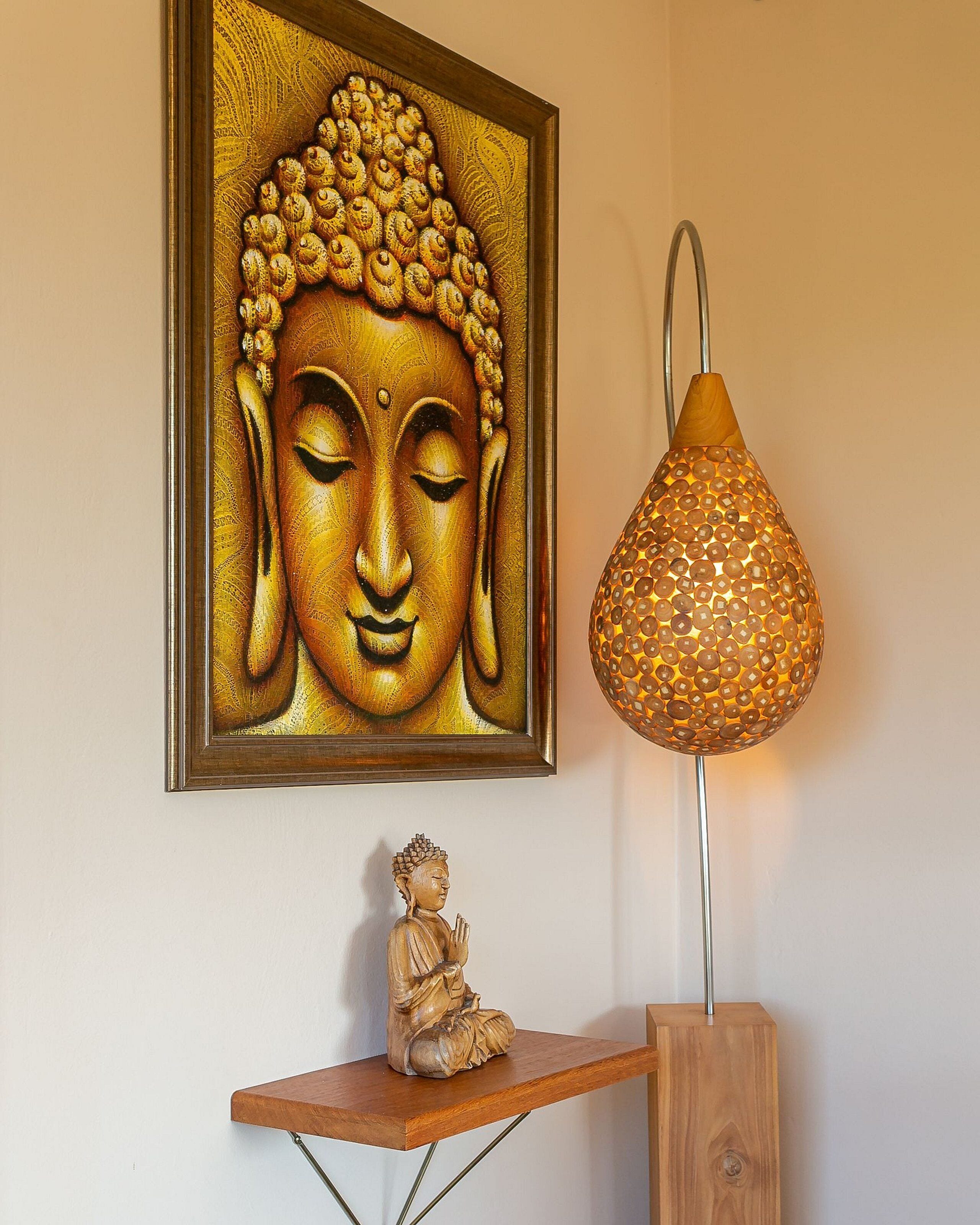 Una estatua de Buda, un cuadro enmarco con Buda y una lámpara decorativa sobre un estante de madera frente a una pared lisa.