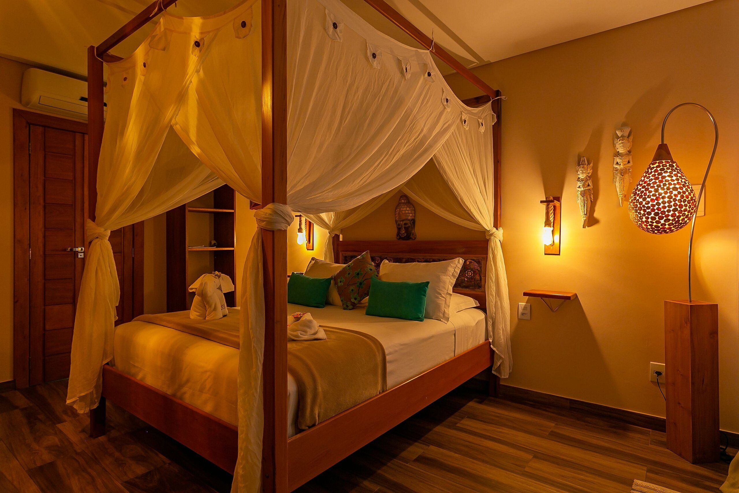 A four-poster bed with a canopy, a wooden floor, a bedside table with a lamp, and wall-mounted artwork.