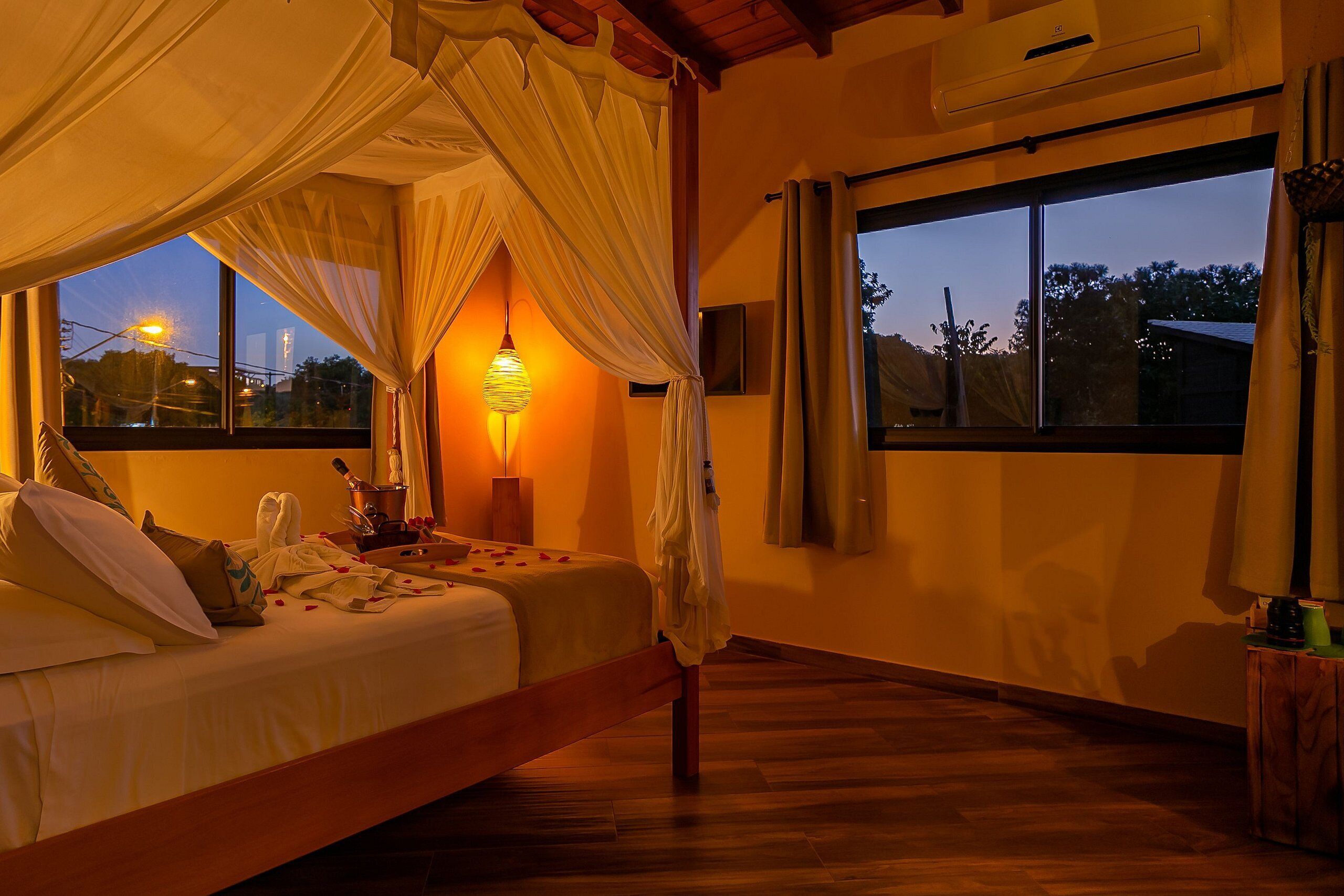 Quarto com cama, luminária de cabeceira e vista para o exterior ao entardecer.