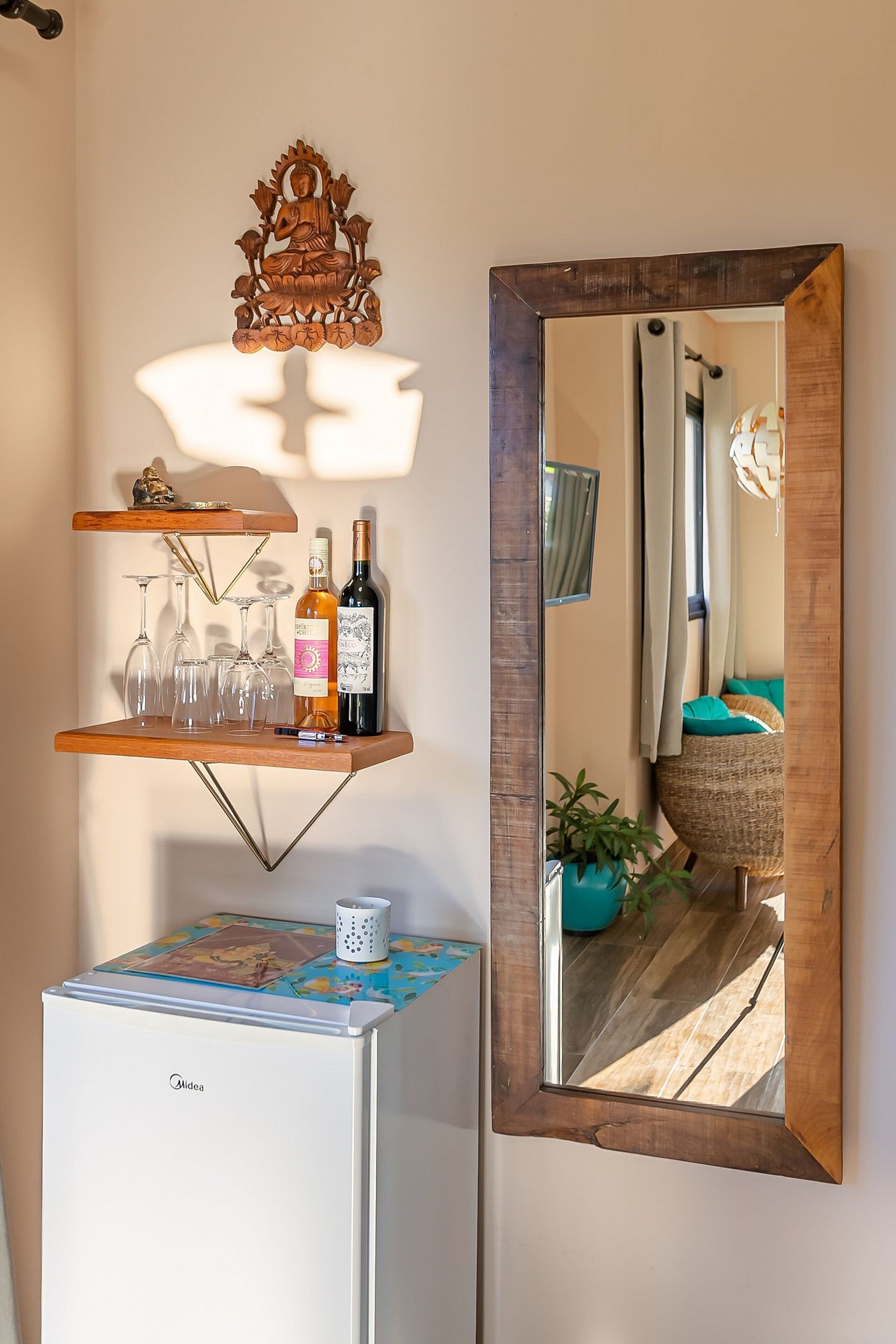 A compact living space with a wooden shelf holding wine glasses and bottles, a white refrigerator, and a mirror reflecting the room.
