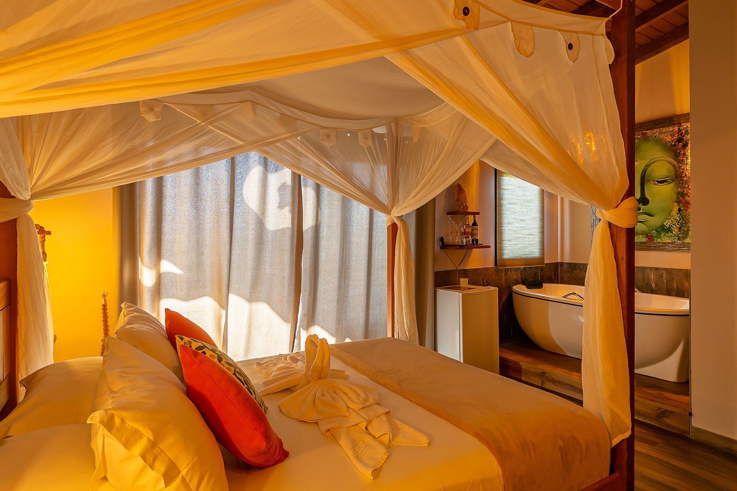 Habitación con cama bajo un dosel, almohada roja y toalla sobre la cama. Se ve una ventana con cortinas, un lavamanos y un espejo al fondo.