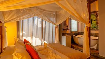 Habitación con cama bajo un dosel, almohada roja y toalla sobre la cama. Se ve una ventana con cortinas, un lavamanos y un espejo al fondo.
