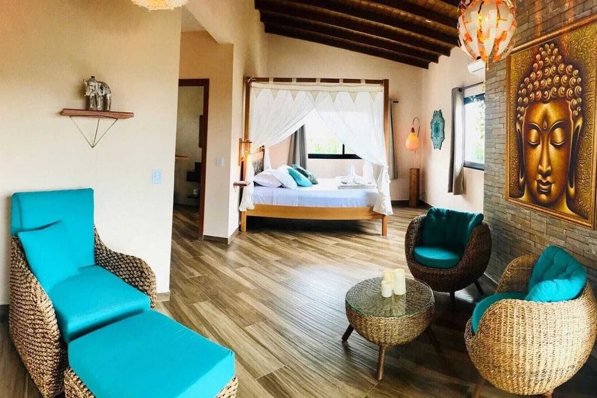 A room with a bed, two chairs, a small table, and a Buddha painting.