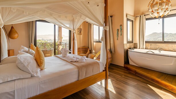 A bedroom with a canopy bed, a bathtub, and large windows offering a view of the hills. - Shiva Boutique Hotel - Praia do Rosa (Imbituba)