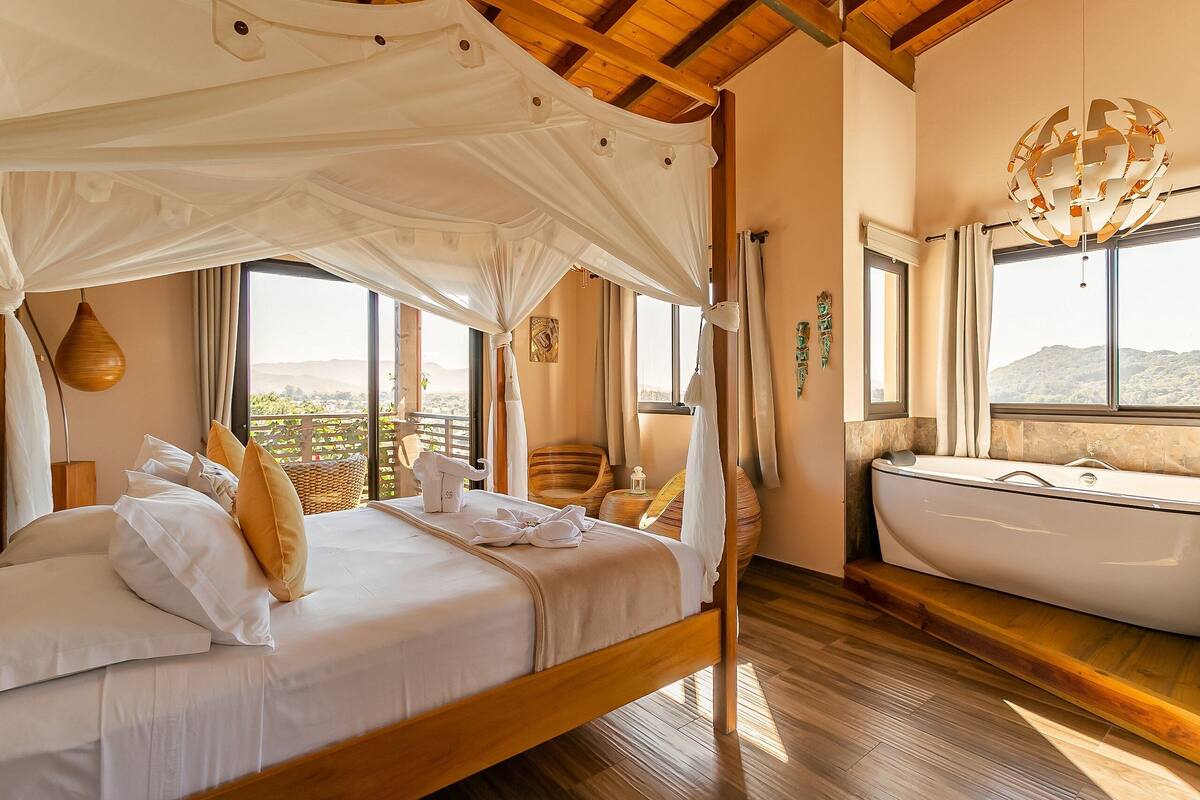 A bedroom with a canopy bed, a bathtub, and large windows offering a view of the hills.