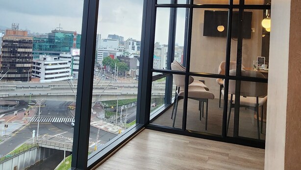 A modern hotel room with large floor-to-ceiling windows, a dining area with a table and chairs, and a flat-screen TV mounted on the wall.