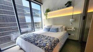 A modern bedroom with a large bed, a bedside telephone, a plant, and a view of the outside balcony.