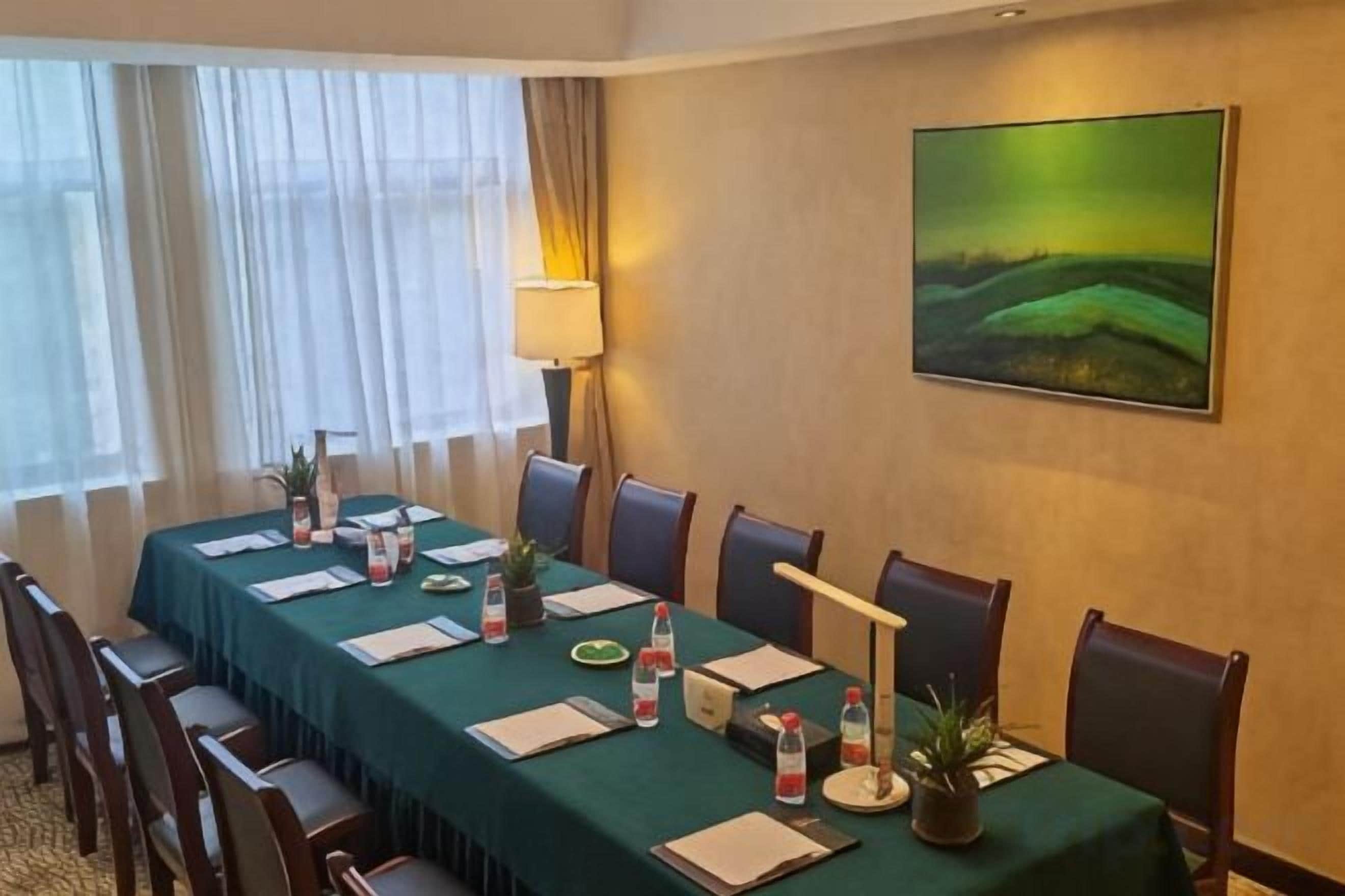 Un salón de conferencias con una larga mesa, sillas, una lámpara, un cuadro y una ventana con cortinas.