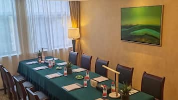 Un salón de conferencias con una larga mesa, sillas, una lámpara, un cuadro y una ventana con cortinas.