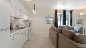 A modern kitchen with white cabinets, a marble countertop, and pendant lighting. There is a beige sofa in the living area with a view of the outdoors.