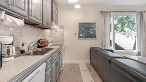 A modern kitchen with grey cabinets, a built-in oven, and a coffee maker. There is a window with curtains and a view of trees outside.