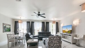 Een moderne woonkamer met een plafondventilator, een flatscreen-tv, een eethoek met een tafel en stoelen, en een fauteuil met een patroon.