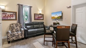 Ein Wohnzimmer mit grauem Sofa, einem gemusterten Sessel, einem Esstisch mit Stühlen und zwei Wandleuchten.