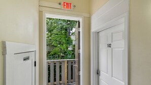 Ein Innenbereich mit einem Flur, einem Ausgangsschild, einer Tür mit der Aufschrift '102' und einem Blick auf einen Balkon mit Geländer und Bepflanzung.