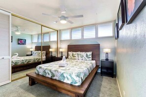 Quarto com um espelho grande, cama com colcha de tema tropical, um banco de madeira e um ventilador de teto.