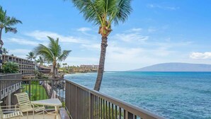 A balcony with a table and chairs, overlooking a beach with palm trees and a clear view of the ocean.