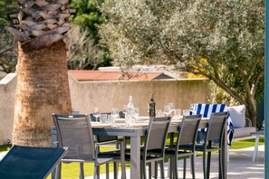 Zona pranzo all'aperto con tavolo imbandito per quattro persone, circondato da sedie e un albero a sinistra.