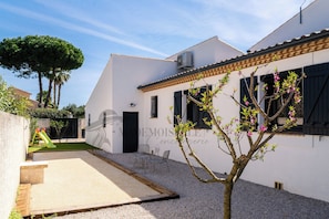 A white building with a black door, a small garden area with a tree and a bench, and a clear sky. - Villa Margot, stunning pool, beach 500 m (Agde)