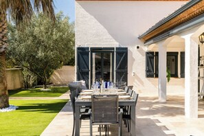 An outdoor patio with a table set for dining, surrounded by a white wall and a garden with olive trees. - Villa Margot, stunning pool, beach 500 m (Agde)