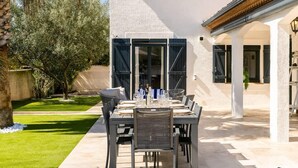 An outdoor dining area with a table set for six, surrounded by chairs, in a courtyard with a white building and a palm tree. - Villa Margot, stunning pool, beach 500 m (Agde)