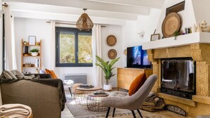 A living room with a sofa, armchair, coffee table, and fireplace. There is a television on a stand, a bookshelf, and a window with curtains. - Villa Margot, stunning pool, beach 500 m (Agde)