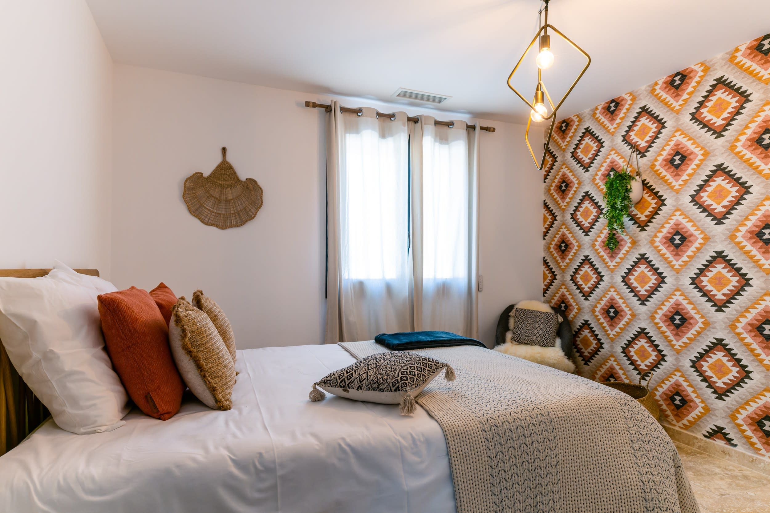 A bedroom with a bed, pillows, a hanging light fixture, a window with curtains, and a patterned wall.