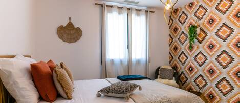 A bedroom with a bed, pillows, a hanging light fixture, a window with curtains, and a patterned wall.
