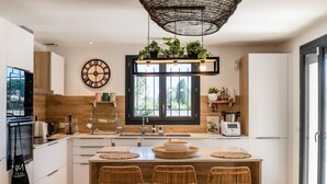 A modern kitchen with a central island, wooden cabinetry, a large clock, and a hanging light fixture. - Villa Margot, stunning pool, beach 500 m (Agde)