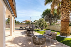 An outdoor patio with a dining table, chairs, and a sofa. There is a palm tree and a hedge in the background. - Villa Margot, stunning pool, beach 500 m (Agde)