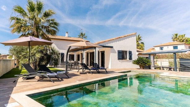 A swimming pool with lounge chairs and umbrellas, surrounded by palm trees and a white building with a tiled roof. - Villa Margot, stunning pool, beach 500 m (Agde)