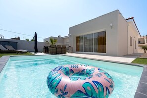 A modern pool with a pink inflatable ring, a covered patio area, and a building with large windows and a flat roof. - Villa Juxta Maré, Beach 500 m Away (Sérignan)