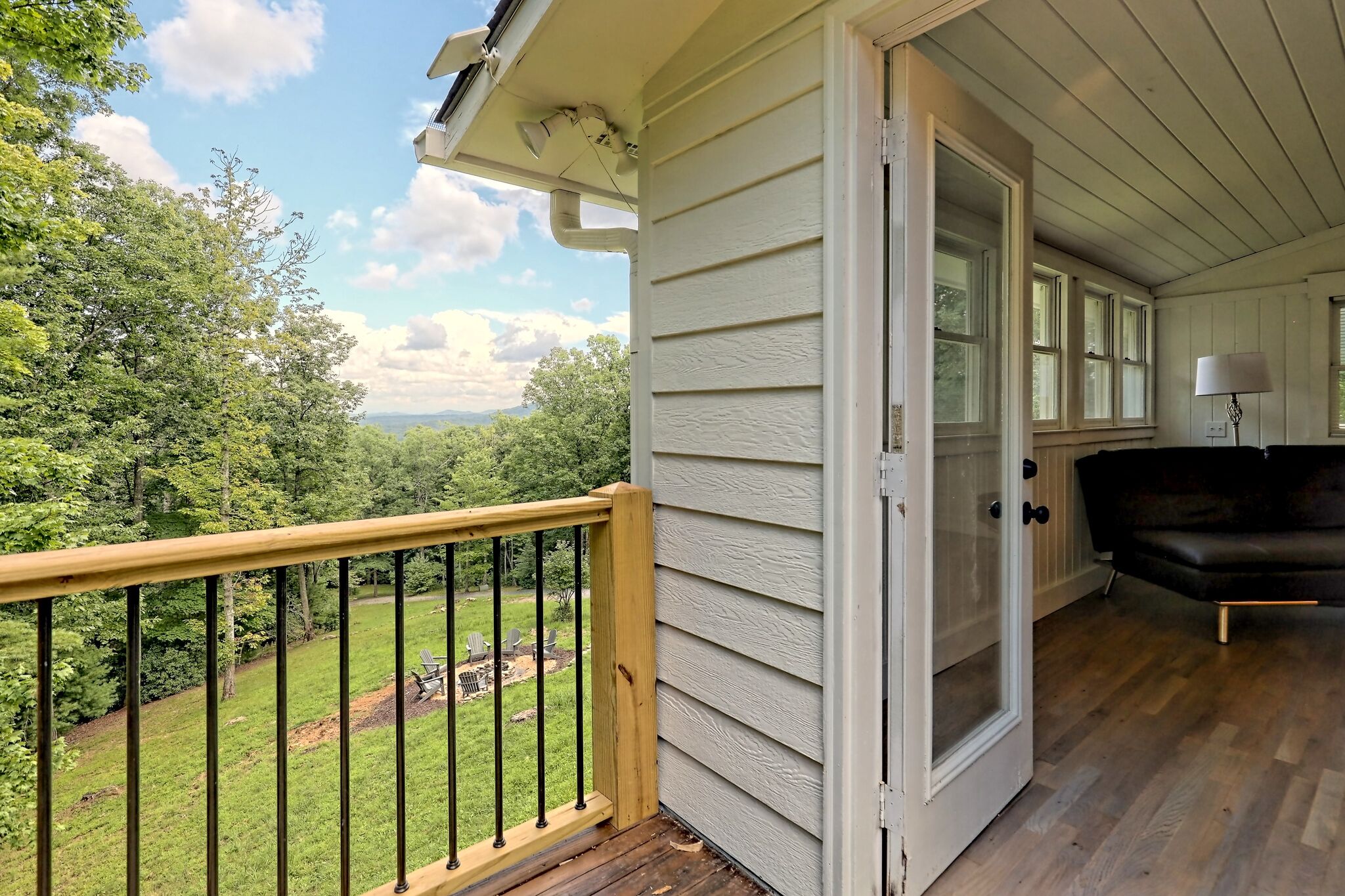 Eine Holzterrasse mit Geländer, eine Glastür zu einem Raum mit einem schwarzen Ledersofa und einer Lampe, und Blick auf eine grüne Landschaft mit Bäumen und blauem Himmel.
