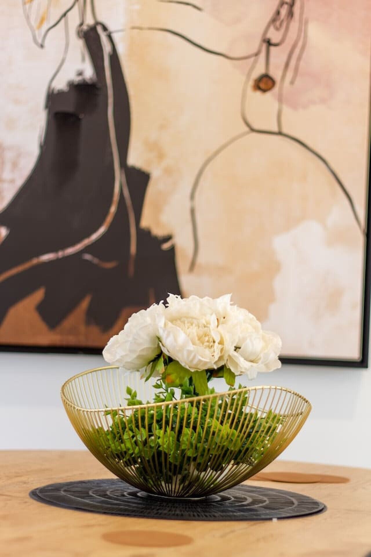 Un tazón decorativo con flores blancas y follaje verde sobre una superficie de madera, con una pintura de fondo.