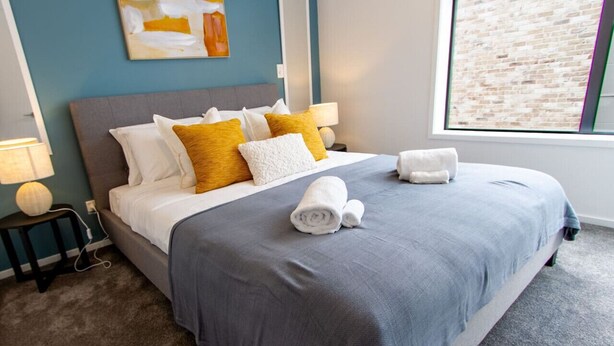 Una cama bien tendida con colcha gris y almohadas blancas, una lámpara en una mesita de noche y una ventana con vista a un muro de ladrillo.