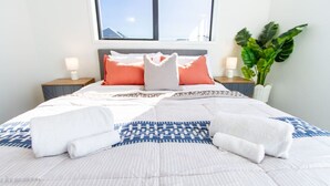 A neatly made bed with patterned bedding, a large window with a view, and a potted plant on the nightstand. - Gateway to Hagley Park - 2 Bed w/Carpark (Christchurch)