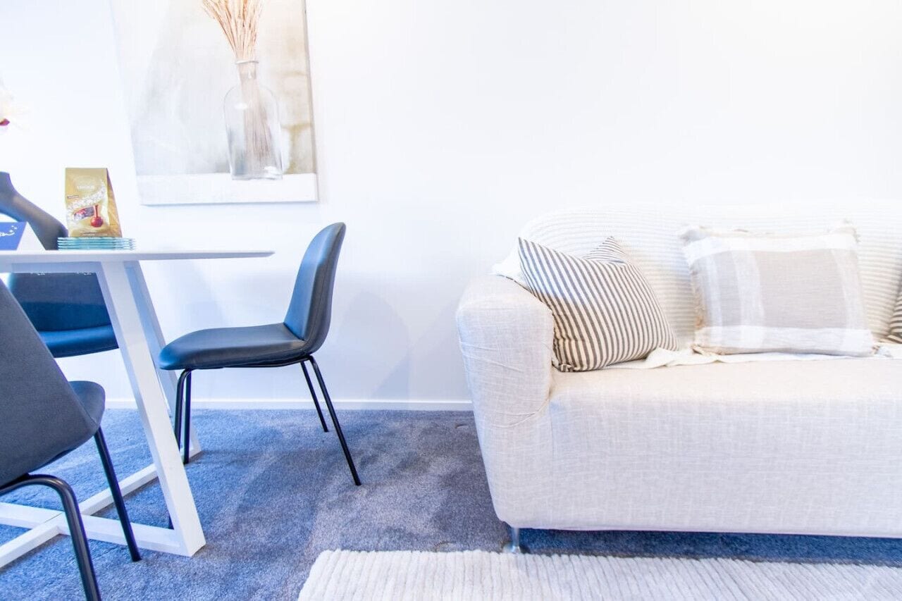 Una sala de estar moderna con un sofá blanco, almohadas a rayas, una silla azul y una mesita con un jarrón y un marco de fotos.