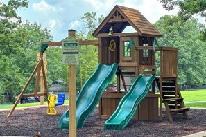Explore the community playground, featuring a play structure with two slides and a swing set, surrounded by a lush green area and trees.