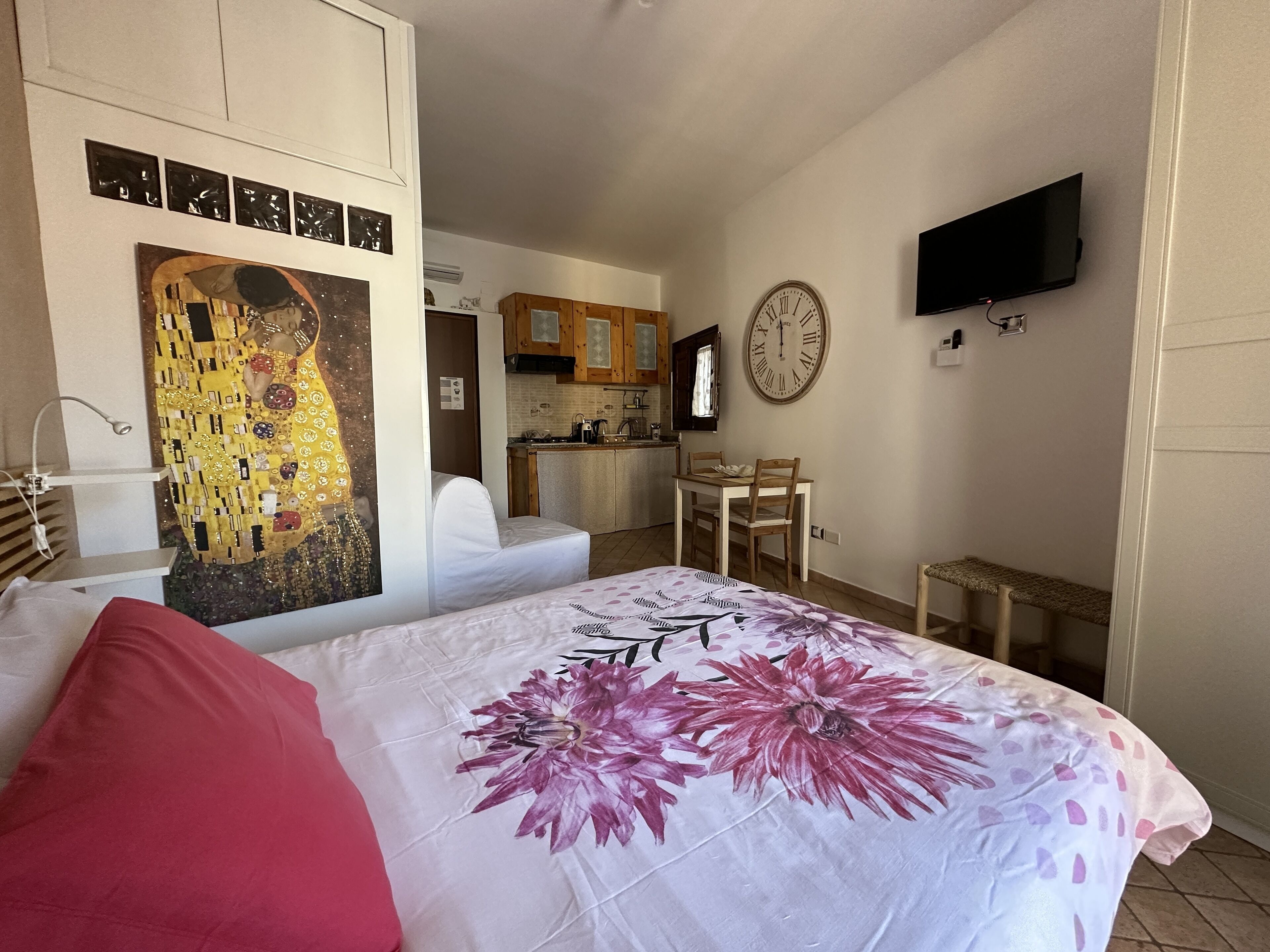 Un dormitorio con cama, un cuadro, un reloj, un televisor y una kitchenette al fondo.