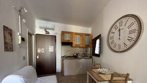 Una stanza compatta con un letto, un tavolo di legno con sedie, un angolo cottura con mobili in legno e una grande parete orologio.