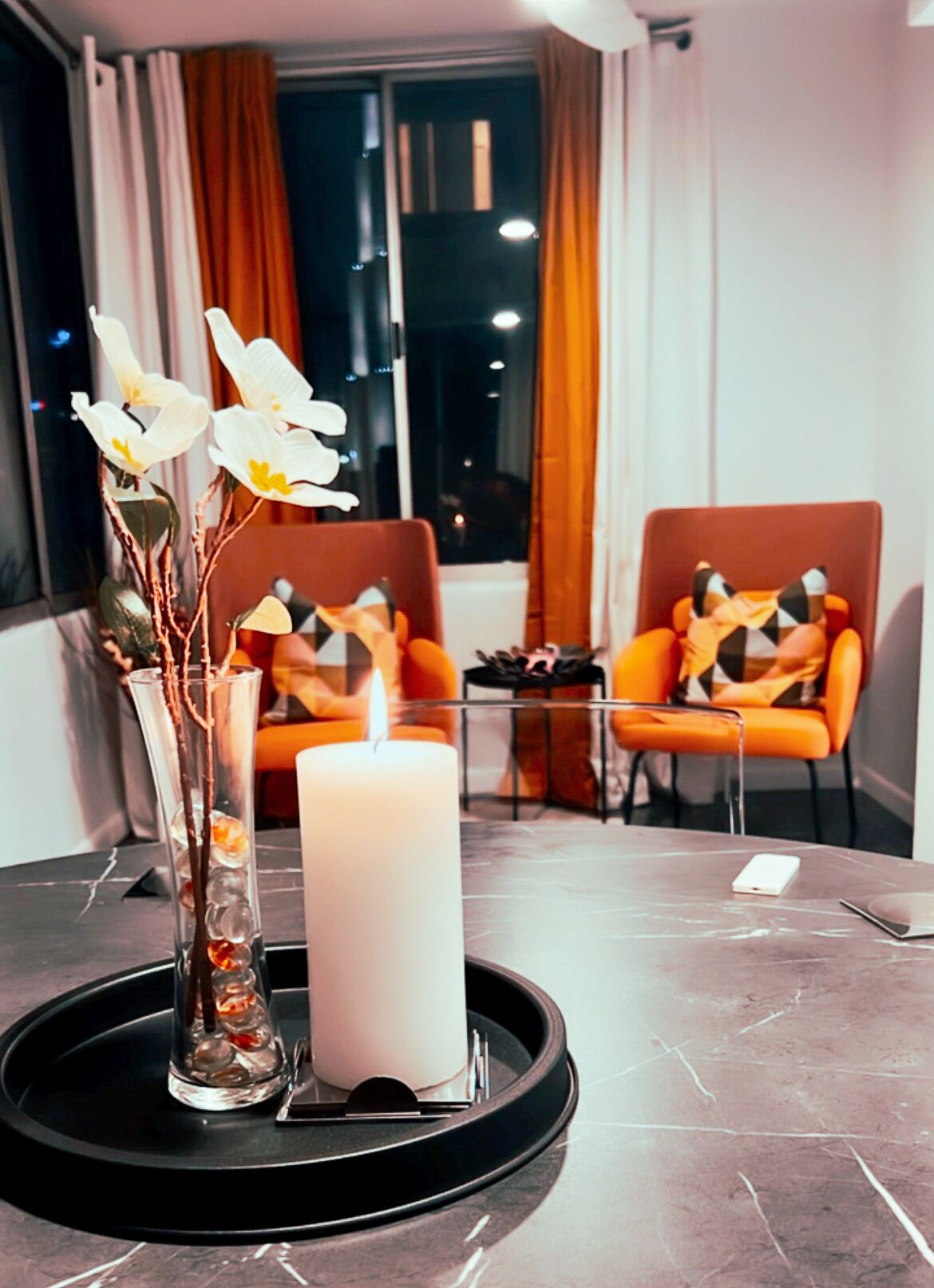 Ein modernes Zimmer mit einem runden Tisch, einer Kerze und einer Vase mit Blumen. Es gibt zwei orangefarbene Stühle mit Katzenmuster.