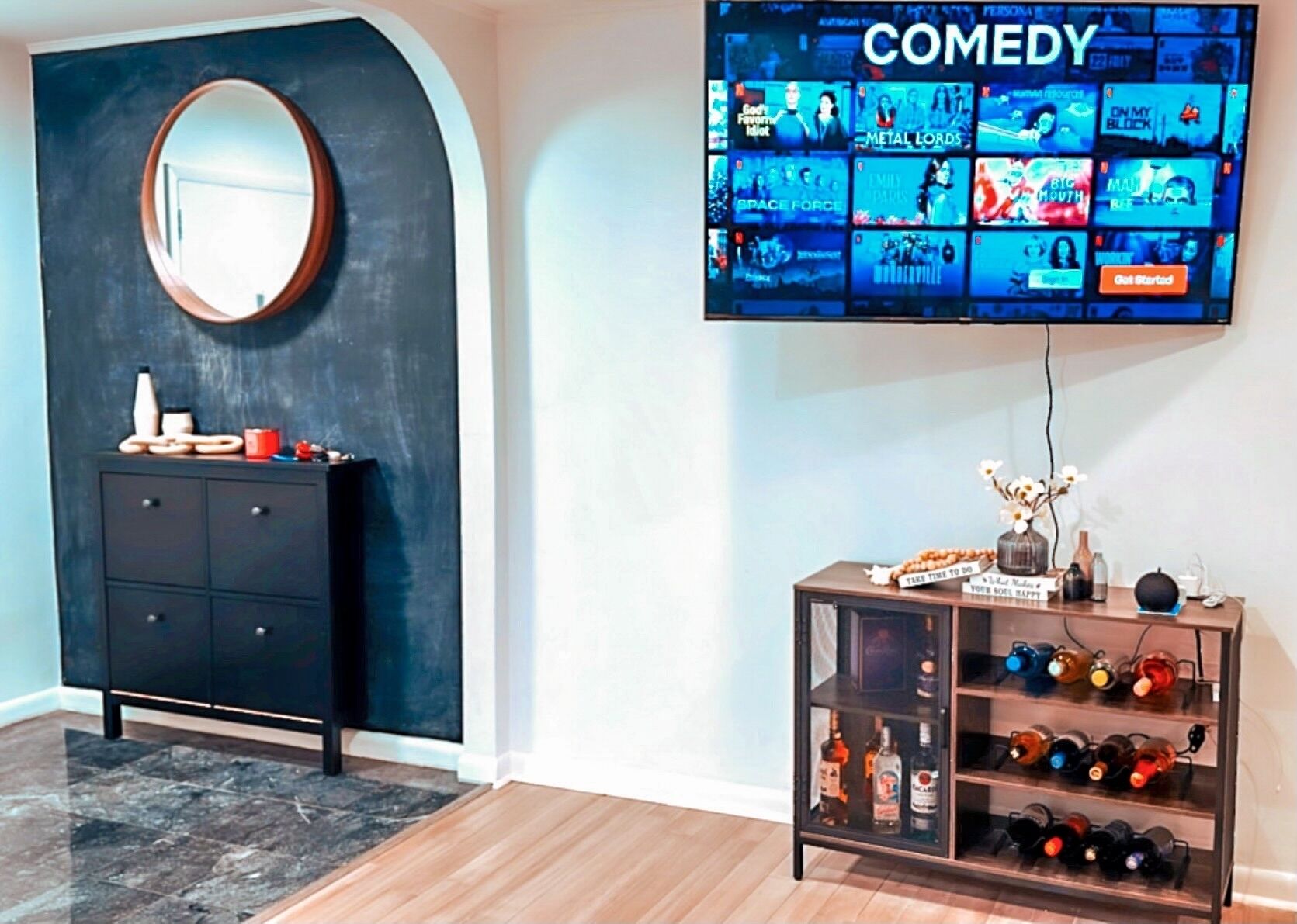 A room with a black curved wall, a round mirror, a black cabinet with decorative items, a TV displaying a comedy channel, and a wine rack with bottles.