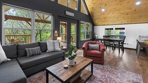 Ein großzügiger Wohnbereich mit hohen Decken, großen Fenstern, einem dunkelgrauen Sofa, einem roten Sessel, einem Holztisch und einem Esstisch mit Stühlen.