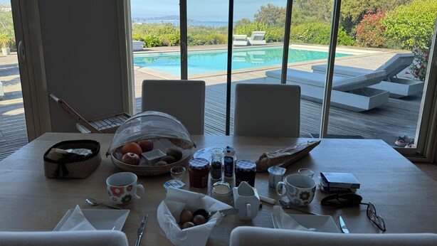 Una mesa de comedor con alimentos para el desayuno y vistas a una piscina y sillones de descanso.