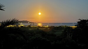 Vista al atardecer desde un balcón con vistas a una playa, palmeras y un horizonte urbano lejano.
