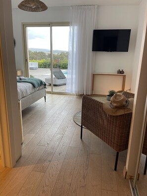 A modern hotel room with a bed, a wicker chair, a TV, and a view of a pool and mountains.