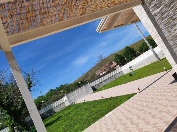 A covered outdoor area with a view of a building in the background, a paved walkway, and a grassy area.