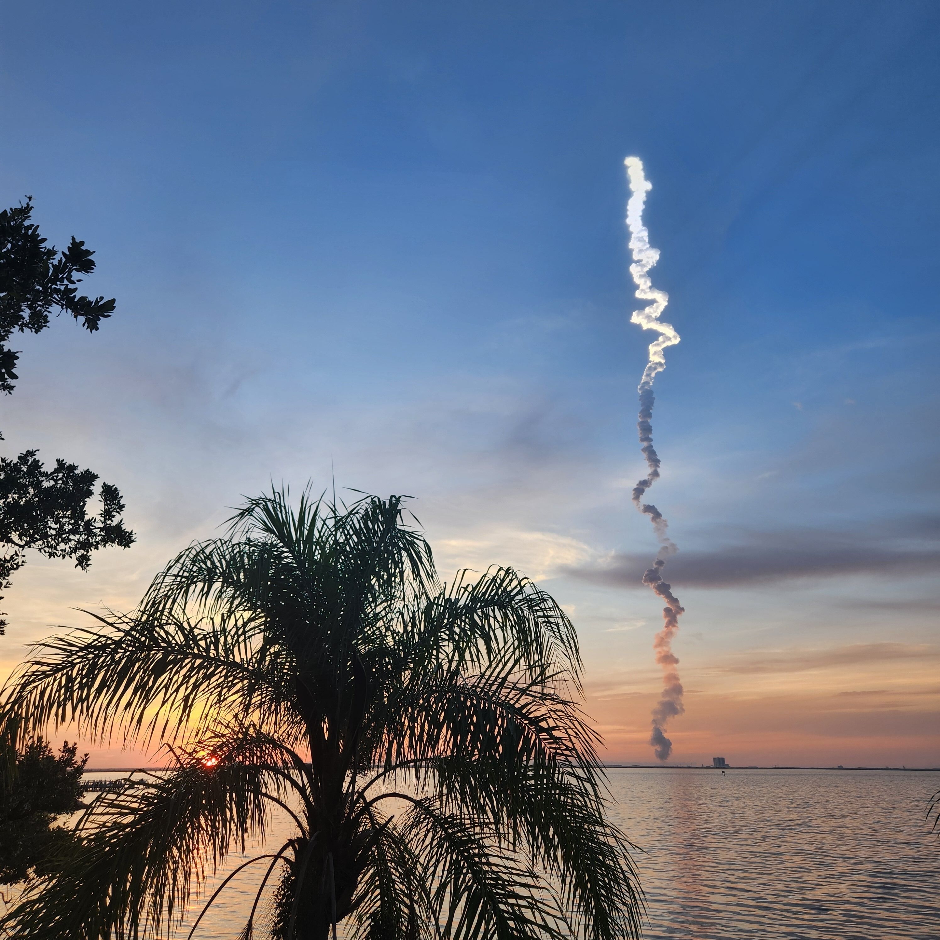 A sunset over a body of water with a palm tree in the foreground and a smoke trail in the sky.