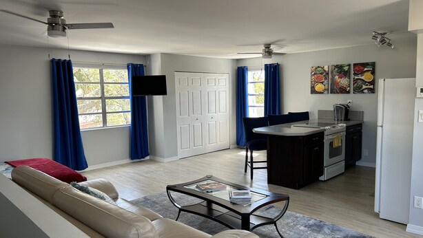 Una sala de estar moderna con un sofá, una mesa de centro y un televisor. Hay una zona de cocina con un refrigerador, estufa y mesa de comedor.