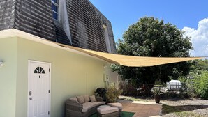 Un patio cubierto con un conjunto de asientos acolchados, una puerta con la etiqueta 'B' y un árbol grande al fondo.