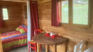 Une cabane chaleureuse avec un intérieur en bois, un lit recouvert d’une couverture colorée, une table à manger avec des chaises et une fenêtre avec des rideaux.